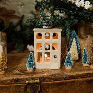 May include: A white ceramic house-shaped candle holder with a window and a wreath on the door. The candle holder is lit with a small tea light candle inside. The candle holder is surrounded by miniature artificial pine trees.