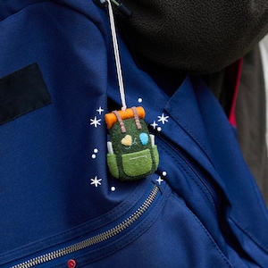 May include: A felt backpack ornament in shades of green, orange, and brown, hanging from a white string. The miniature backpack has a front pocket and is decorated with small details. It is attached to a blue backpack.