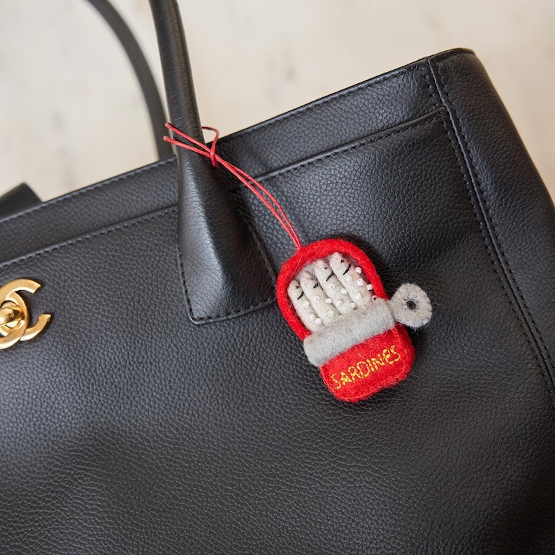 Handmade felt sardine tin bag charm tied to black leather purse, whimsical fish accessory with red can design, quirky seafood gift for food lovers