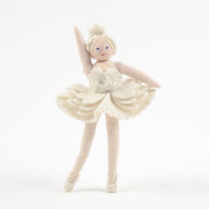 May include: A felt ballerina doll in a cream-colored outfit with a layered tutu skirt, a bodice with subtle detailing, and a small crown on its head. The doll has light skin, blue eyes, and is posed with one arm raised in a dance position.