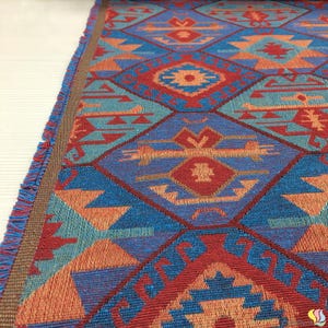 May include: Close-up of a woven textile with a geometric pattern in shades of blue, red, and orange. The design features diamond shapes and stylized tribal motifs. The edges are finished with a brown border and fringe.