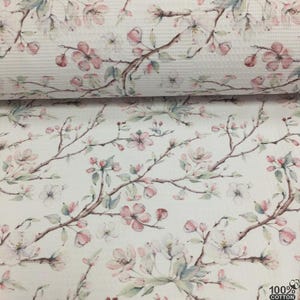 May include: White cotton fabric with a floral pattern featuring pink and white blossoms on brown branches. The design includes green leaves and a textured waffle weave. The fabric is labeled as 100% cotton.
