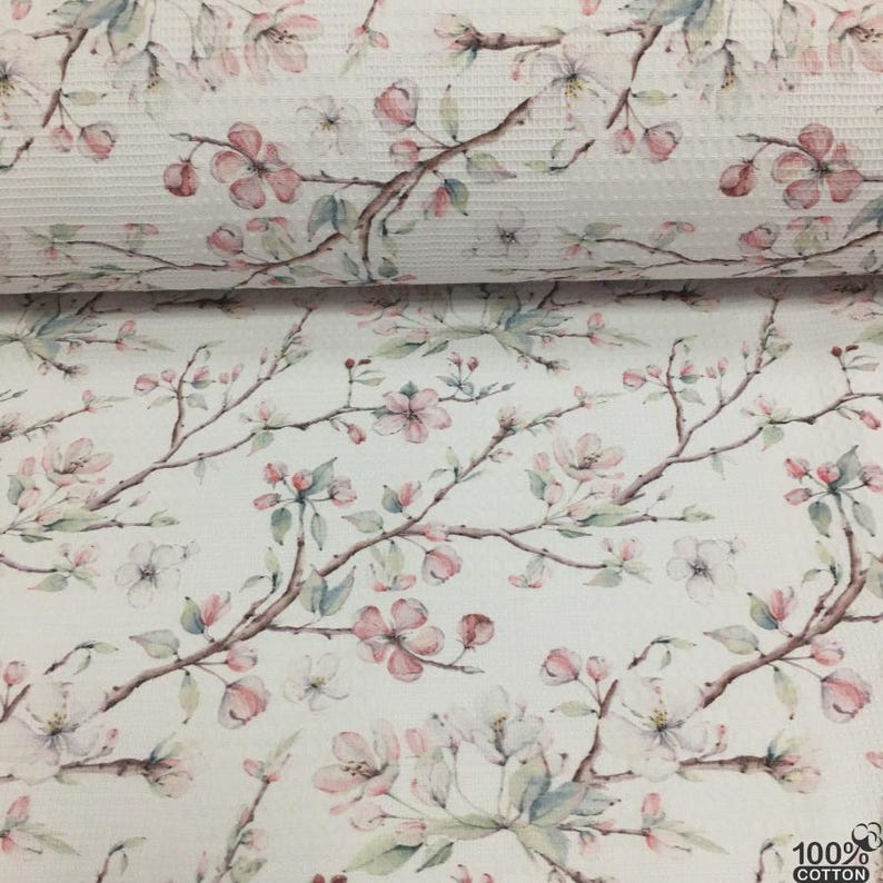 May include: White cotton fabric with a floral pattern featuring pink and white blossoms on brown branches. The design includes green leaves and a textured waffle weave. The fabric is labeled as 100% cotton.