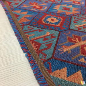 May include: Close-up of a woven textile with a geometric pattern. The design features blue, red, and orange shapes, including diamonds and arrows. The edges are finished with a brown border and blue fringe.