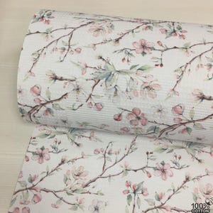 May include: White cotton fabric with a floral pattern featuring pink blossoms and green leaves on brown branches. The fabric is rolled and shows a textured waffle weave. The bottom right corner shows the text "100% COTTON".