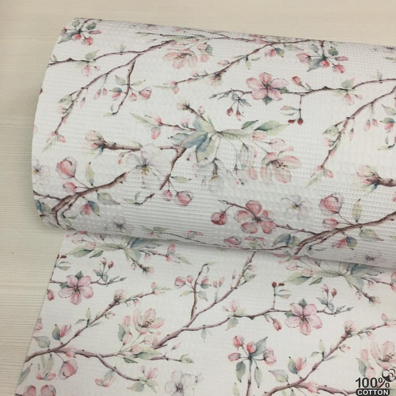 May include: White cotton fabric with a floral pattern featuring pink blossoms and green leaves on brown branches. The fabric is rolled and shows a textured waffle weave. The bottom right corner shows the text "100% COTTON".