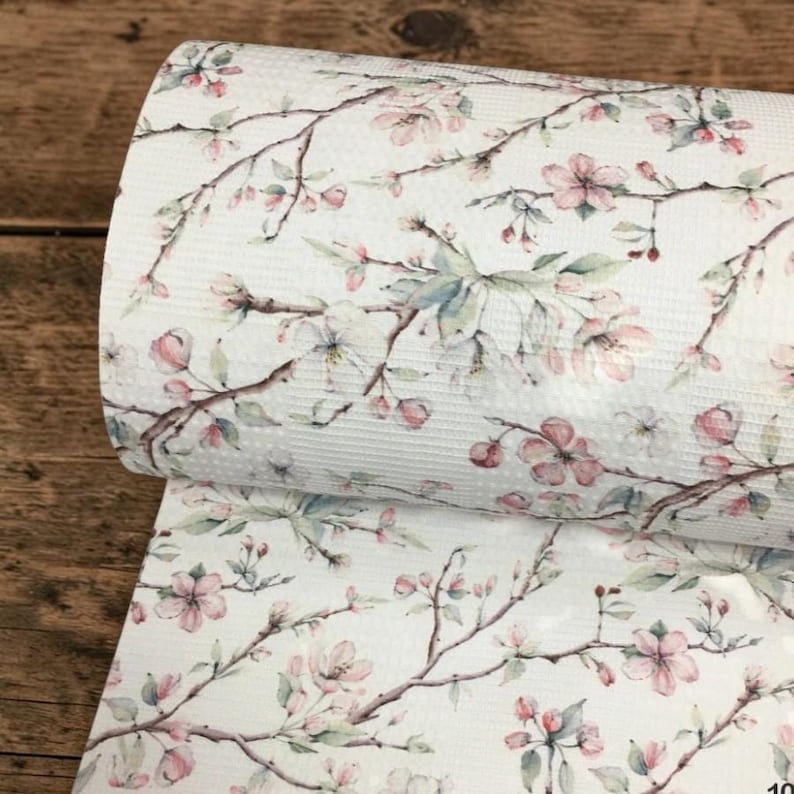 May include: A roll of white fabric with a delicate floral pattern. The design features branches with pink blossoms and green leaves. The fabric has a textured surface, and the overall aesthetic is soft and feminine.