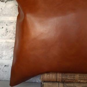May include: A large, brown leather cushion with a smooth surface. The cushion is resting on a stack of vintage books. The background features a white brick wall.