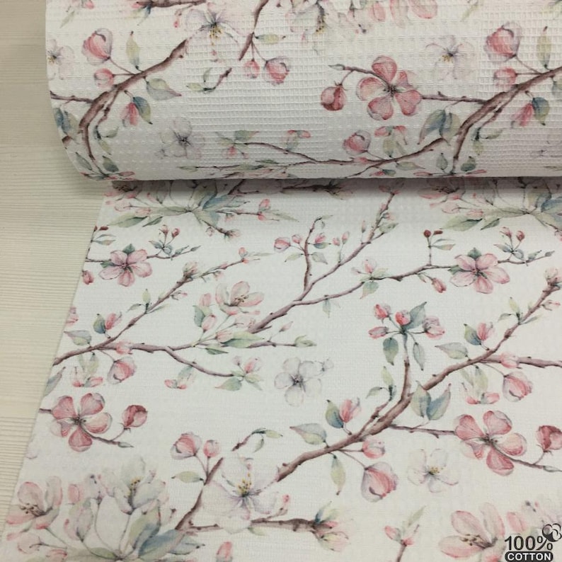 May include: White cotton fabric with a floral pattern featuring pink and white blossoms, green leaves, and brown branches. The fabric is rolled up, showing the textured weave. The bottom of the fabric is spread out, displaying the full floral design.