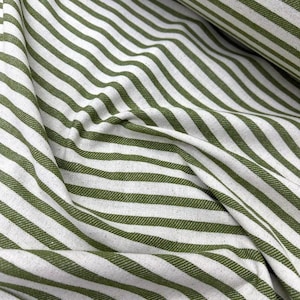 Pode incluir: Close-up de um tecido com um padrão repetitivo de listras verde-oliva e brancas. O tecido é dobrado e drapeado, mostrando a textura e a direção das listras. As listras são espaçadas uniformemente e correm na diagonal.