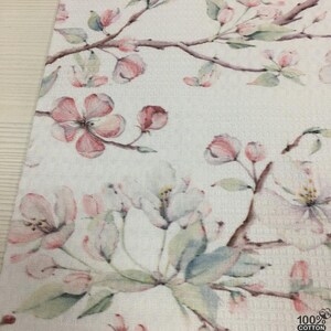 May include: White floral patterned fabric featuring delicate pink and white blossoms and green leaves on brown branches. The fabric has a textured waffle weave. The lower right corner shows the text "100% COTTON".