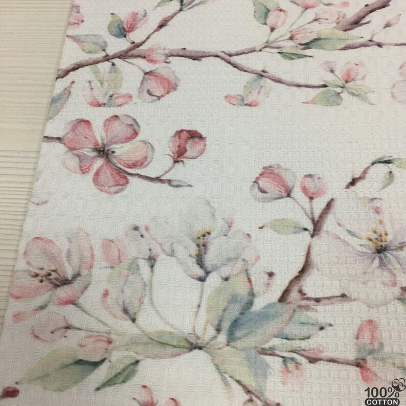 May include: White floral patterned fabric featuring delicate pink and white blossoms and green leaves on brown branches. The fabric has a textured waffle weave. The lower right corner shows the text "100% COTTON".