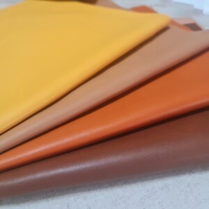 May include: A selection of leather sheets in shades of yellow, tan, orange, and brown. The sheets are stacked, revealing their different colours and textures. The leather has a smooth finish, indicating a quality product.