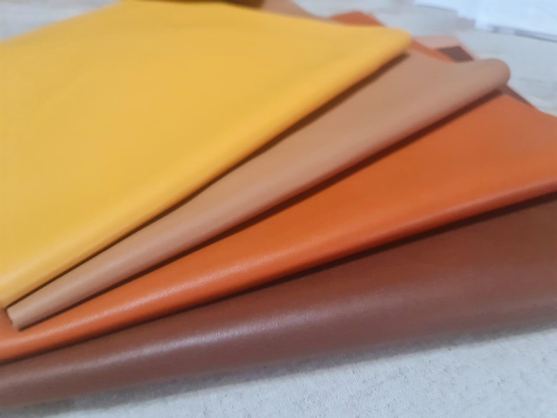 May include: A selection of leather sheets in shades of yellow, tan, orange, and brown. The sheets are stacked, revealing their different colours and textures. The leather has a smooth finish, indicating a quality product.