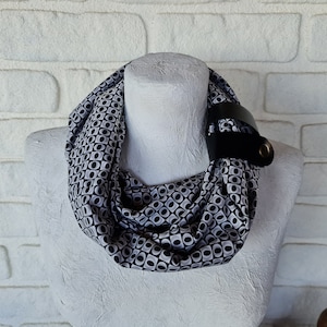 May include: A black and white patterned infinity scarf with a geometric design. The scarf is secured with a black leather strap and a gold-colored metal eyelet. The scarf is displayed on a white mannequin head against a white brick wall.