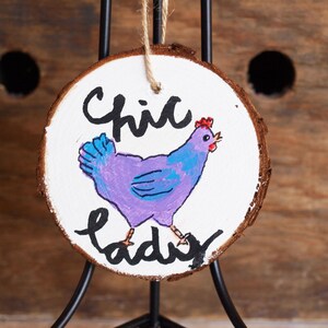 Puede incluir: Un adorno redondo de madera con fondo blanco y un pollo morado pintado en él. El pollo tiene alas azules y una cresta roja. Las palabras "Chic Lady" están escritas en negro debajo del pollo.