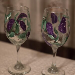 May include: Two clear glass wine glasses featuring hand-painted purple floral clusters and green leaves. The stems are slender and the bases are circular. The painted designs wrap around the bowls of the glasses, showcasing detailed brushstrokes in shades of violet and sage green on the transparent surfaces.