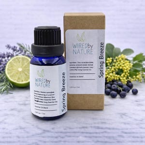 May include: A blue glass essential oil bottle with a black cap and a matching cardboard box. The label reads "Wired by Nature Spring Breeze Essential Oil Blend". The image includes a lime, lavender, and berries.