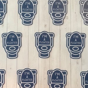 May include: Nine blue and white toilet-shaped signs with the text "I'm a Floater" on a white wood background.