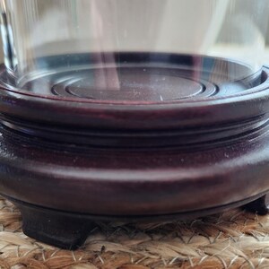 May include: A dark brown, wooden display stand with a circular top and a rounded base. The stand has a tiered design and is supported by three small feet. The stand is designed to hold a glass cloche or similar decorative item.