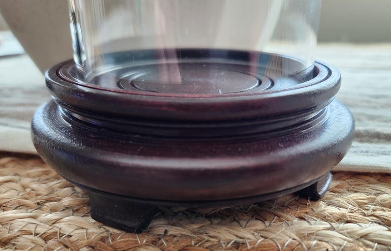 May include: A dark brown, wooden display stand with a circular top and a rounded base. The stand has a tiered design and is supported by three small feet. The stand is designed to hold a glass cloche or similar decorative item.