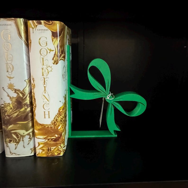 Puede incluir: Un sujetalibros verde en forma de lazo se encuentra entre dos libros de tapa dura con letras y detalles dorados. El sujetalibros est&aacute; hecho de un material s&oacute;lido y tiene un dise&ntilde;o sencillo. Los libros est&aacute;n en una estanter&iacute;a negra.
