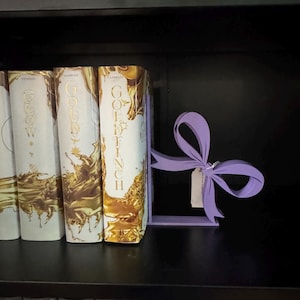 Puede incluir: Un sujetalibros decorativo en forma de lazo lavanda sostiene una fila de libros con letras y detalles dorados. El sujetalibros tiene una peque&ntilde;a etiqueta adjunta. Los libros est&aacute;n en una estanter&iacute;a negra.