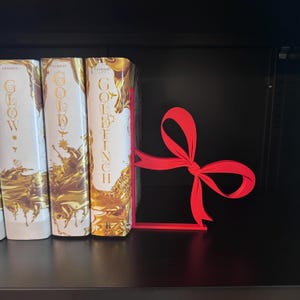 Puede incluir: Un sujetalibros rojo en forma de lazo se encuentra en una estanter&iacute;a negra junto a tres libros con cubiertas doradas y blancas. El sujetalibros est&aacute; hecho de un material s&oacute;lido y tiene un dise&ntilde;o simple y elegante.