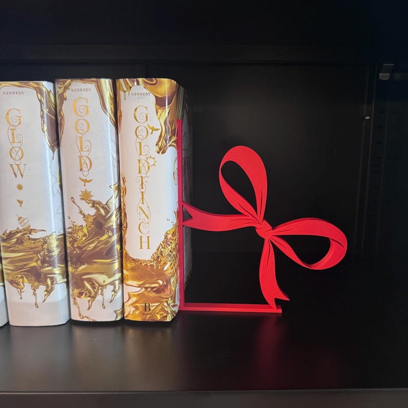 Puede incluir: Un sujetalibros rojo en forma de lazo se encuentra en una estanter&iacute;a negra junto a tres libros con cubiertas doradas y blancas. El sujetalibros est&aacute; hecho de un material s&oacute;lido y tiene un dise&ntilde;o simple y elegante.