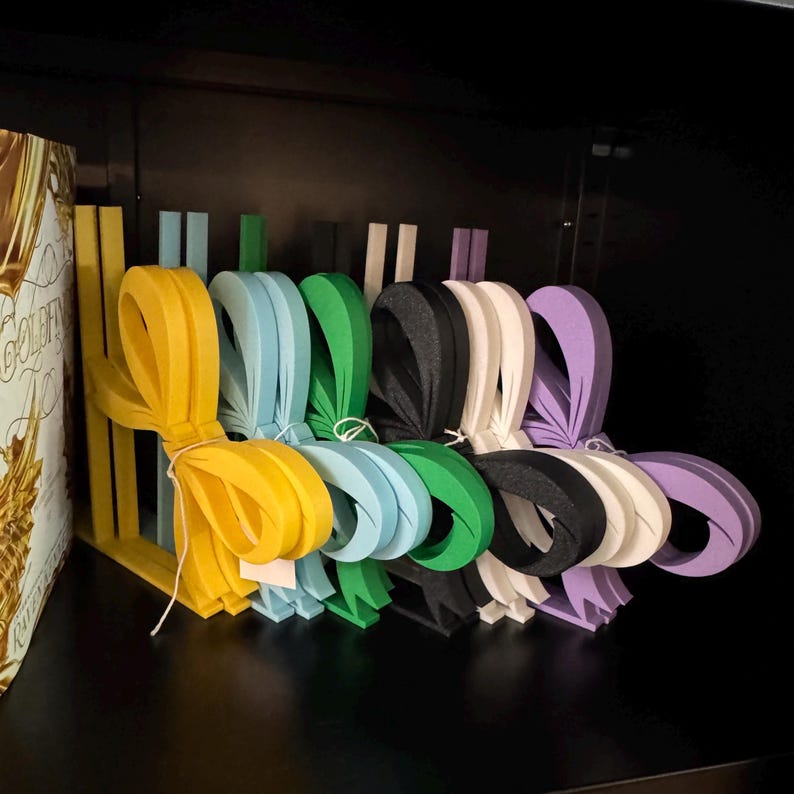 Puede incluir: Una colecci&oacute;n de lazos de papel coloridos en varios tonos, incluyendo amarillo, azul, verde, negro, blanco y morado. Cada lazo est&aacute; atado con una cuerda blanca y dispuesto en fila, sugiriendo un tema de manualidades o envoltorio de regalo.