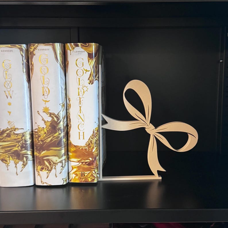 Puede incluir: Un sujetalibros de madera en forma de lazo, junto a tres libros con cubiertas doradas y blancas. El sujetalibros es de color marr&oacute;n claro y tiene una base plana. Los libros tienen letras doradas y un dise&ntilde;o l&iacute;quido dorado.