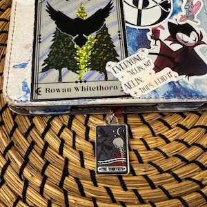May include: A close-up of a tarot card bookmark with a dark blue background and a white figure with long hair. The card is labeled "The Thirteen" and has the text "Until the darkness drives me in." The bookmark is hanging from a white notebook with stickers that say "Everyone: "Aelin, no" and "Aelin: Oops, I did it."