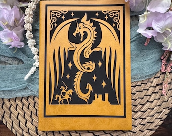 May include: A tan leather journal with a black dragon design. The dragon is centered on the cover, with a knight on horseback and a castle below. The journal is rectangular and has a border.