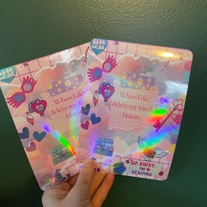 May include: Two pink and purple holographic bookmarks with a whimsical design featuring a pair of glasses, hearts, and the text "When I die, delete my kindle history" and "Go away, I'm reading".