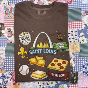 May include: Brown t-shirt with a graphic of Saint Louis landmarks and food icons. The design includes the Gateway Arch, a fleur-de-lis, and a baseball. The text "SAINT LOUIS" and "THE LOU" are also featured.