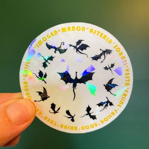 K&ouml;nnte beinhalten: Ein wei&szlig;er, schimmernder Aufkleber mit einem Kreis aus schwarzen Drachen-Silhouetten und goldenem Text am Rand. Der Text lautet "Imogen, Manon, Asterin, Sorrel, Vesta, Caline, Eda, Briar, Thren, Krinnea, Gilsea".
