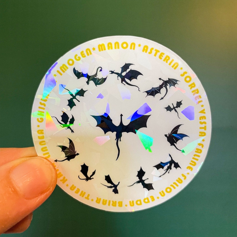 K&ouml;nnte beinhalten: Ein wei&szlig;er, schimmernder Aufkleber mit einem Kreis aus schwarzen Drachen-Silhouetten und goldenem Text am Rand. Der Text lautet "Imogen, Manon, Asterin, Sorrel, Vesta, Caline, Eda, Briar, Thren, Krinnea, Gilsea".