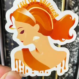 May include: A sticker with a woman with orange hair and a gold crown. The text "I see you, Goldenfinch" is written around the crown.