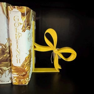 Puede incluir: Un sujetalibros con forma de lazo amarillo brillante sostiene dos libros. El sujetalibros es de madera y tiene un dise&ntilde;o sencillo. Los libros tienen cubiertas doradas y blancas, con el t&iacute;tulo "Goldfinch" visible.