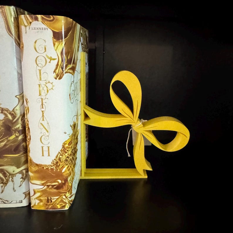 Puede incluir: Un sujetalibros con forma de lazo amarillo brillante sostiene dos libros. El sujetalibros es de madera y tiene un dise&ntilde;o sencillo. Los libros tienen cubiertas doradas y blancas, con el t&iacute;tulo "Goldfinch" visible.