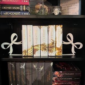 Puede incluir: Una estanter&iacute;a negra con sujetalibros blancos en forma de lazo. La estanter&iacute;a contiene varios libros con cubiertas blancas y doradas, incluyendo t&iacute;tulos como "Glow" y "Goldfinch". Se ven libros adicionales con cubiertas oscuras debajo. Una vela decorativa y flores secas est&aacute;n en la estanter&iacute;a superior.