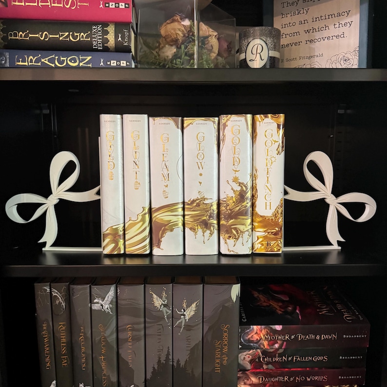 Puede incluir: Una estanter&iacute;a negra con sujetalibros blancos en forma de lazo. La estanter&iacute;a contiene varios libros con cubiertas blancas y doradas, incluyendo t&iacute;tulos como "Glow" y "Goldfinch". Se ven libros adicionales con cubiertas oscuras debajo. Una vela decorativa y flores secas est&aacute;n en la estanter&iacute;a superior.