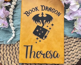 May include: Mustard-colored journal with the words "Book Dragon" and a dragon illustration above a book graphic. The name "Theresa" is written in cursive below the book graphic. A gray ribbon bookmark is visible.
