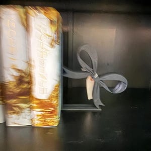 Puede incluir: Un sujetalibros de metal gris con forma de lazo, que sostiene libros con un dise&ntilde;o dorado y blanco. El sujetalibros tiene una peque&ntilde;a etiqueta adjunta. Los libros est&aacute;n sobre una superficie negra.