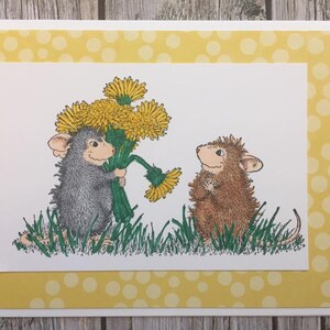 May include: A greeting card with a yellow polka dot border features two mice. One mouse is holding a bouquet of yellow dandelions and the other mouse is looking at the flowers.