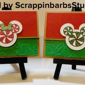 May include: Two handmade Christmas cards featuring a red and green candy cane Mickey Mouse design. The cards are displayed on small wooden easels.
