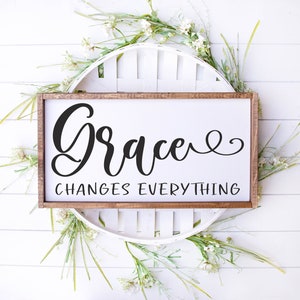 以下が含まれることがあります： 黒い筆記体で「Grace Changes Everything」と書かれた木製の看板。看板は白い花と緑に囲まれている。