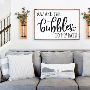 Könnte beinhalten: Ein weißes Schild mit schwarzer Kalligraphie, auf dem "You are the bubbles to my bath" steht. Das Schild hängt an einer weißen Wand über einem grauen Sofa mit Kissen. Zwei hölzerne Wandvasen mit Grünpflanzen befinden sich auf beiden Seiten des Schildes.