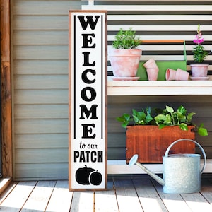 May include: A wooden sign with black lettering that reads "WELCOME to our PATCH" with two black pumpkin silhouettes. The sign is leaning against a white wooden bench with potted plants and gardening tools.