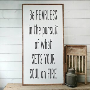 Puede incluir: Un letrero de madera enmarcado con el texto "Be FEARLESS in the pursuit of what SETS YOUR SOUL on FIRE" en letras negras. Una pequeña tabla de lavar de madera y una lata de leche de metal con asa están junto al letrero.